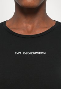 Close-up van een persoon die een zwart shirt draagt met het witte "EA7 Emporio Armani" logo gecentreerd onder de hals.