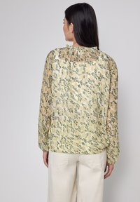 Blumenbluse mit einem hellgelben Hintergrund, die grüne Blätter und kleine rosa Akzente zeigt. Lange Ärmel, die an den Bündchen gerafft sind.
