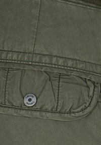 Tejido de prenda en verde oliva con textura acolchada, presenta un bolsillo frontal y un botón plateado circular con la marca "TIMEZONE".