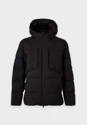 Un homme porte une doudoune noire à capuche, une chemise noire, un pantalon beige à carreaux et des bottes noires sur un fond clair et uni.