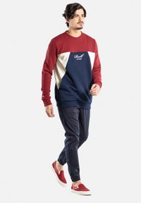 Sudadera bloqueada en rojo, azul marino y crema, con cuello redondo y logo bordado. Combinada con pantalones azul marino y zapatos rojos tipo slip-on.