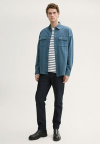 Cămașă button-up de culoare bleu deschis, cu două buzunare pe piept, purtată peste un tricou alb cu dungi navy, asortată cu blugi negri și cizme negre până la gleznă.