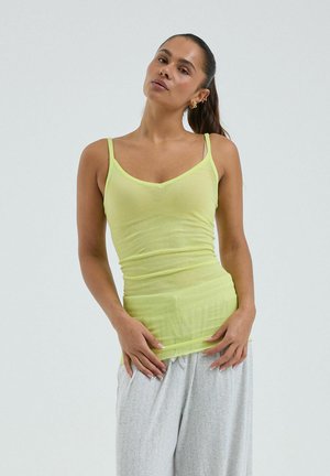 Femme aux longs cheveux foncés portant un haut sans manches vert clair et un pantalon gris clair, debout devant un fond blanc uni.