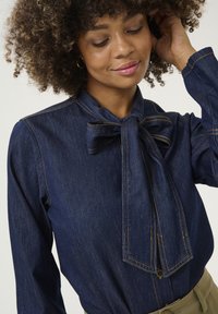Blusa in denim blu scuro con colletto a fiocco, dettagli di cucitura verticale e silhouette aderente. Il tessuto ha una trama liscia.