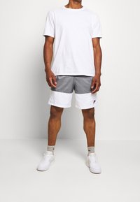 Muž stojící v bílém tričku, šedých a bílých Nike šortkách s barevným blokem, šedých Nike ponožkách a bílých teniskách na světle šedém pozadí.