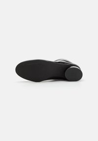 Zapato de cuero negro con un tacón redondeado, superficie lisa y suela texturizada, que presenta un sutil branding y un diseño elegante y moderno.