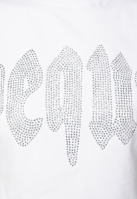 Balta T-kreklu, kas rotāts ar sudraba rhinestones, kas sakārtotas stilizētā burtnīcas dizainā, uzsverot ģeometriskas formas un atstarojošu tekstūru.