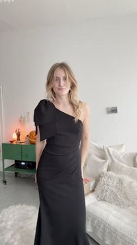 Asymetrické čierne šaty s mašľou na ľavom pleci, priliehavým korzetom a splývavou sukňou s drapériou, vyrobené z hladkej tkaniny.