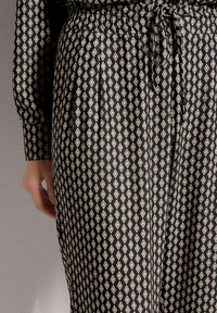 Zwarte stof met een patroon van diamantvormen in beige; elastische tailleband met trekkoord; lange mouwen met knoopmanchet-detail.