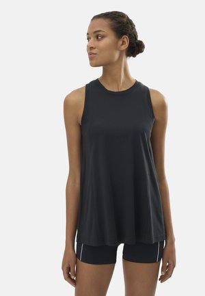 Mujer con una blusa negra sin mangas suelta y pantalones cortos negros ajustados, de pie y mirando hacia su izquierda contra un fondo liso.