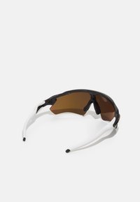 Lunettes de sport avec une monture noire et blanche, verres teintés bruns, design aérodynamique et grips texturés sur le nez pour le confort.