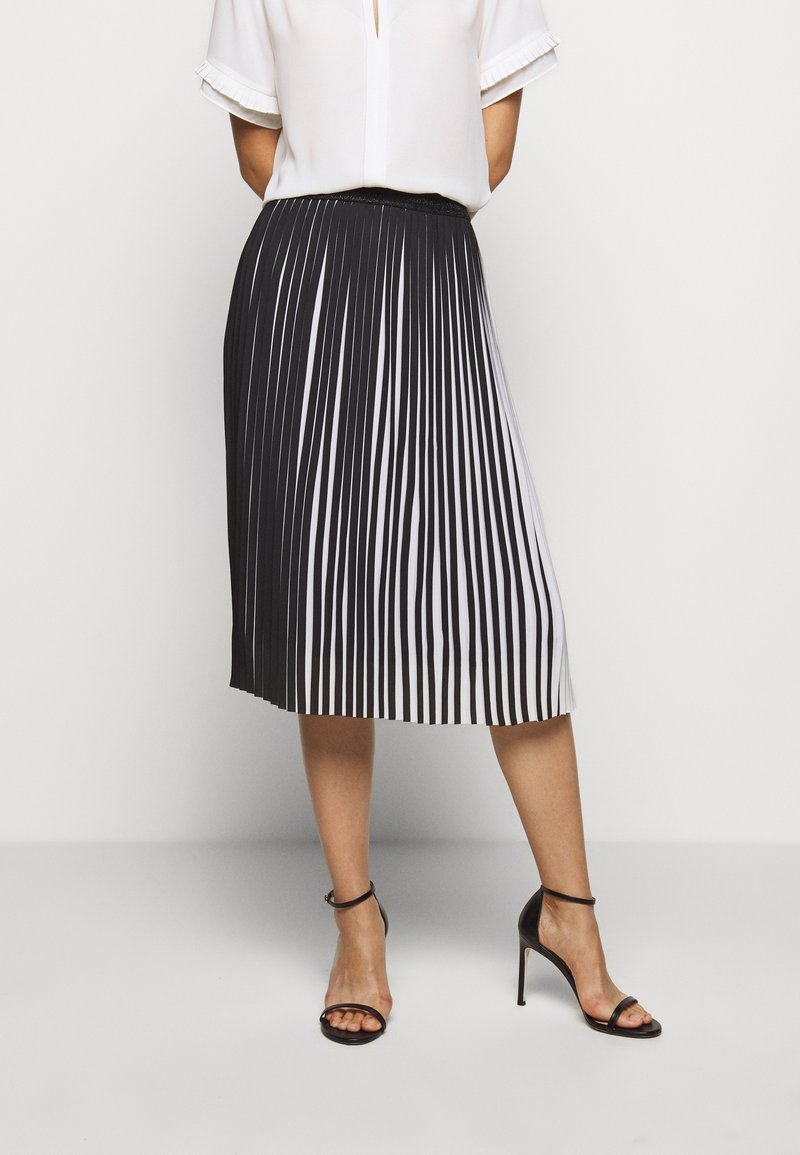 Femme portant une jupe mi-longue plissée noir et blanc, un blouson blanc à manches courtes et des sandales à talons noirs, se tenant devant un fond blanc.