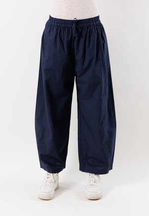 Persona che indossa pantaloni blu navy larghi con vita elastica e sneakers bianche, in piedi contro uno sfondo semplice.
