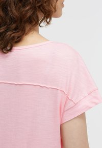 Woman with curly brown hair wears a light pink short-sleeve shirt with a horizontal seam across the back and rolled cuff sleeves.