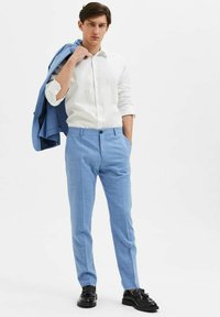 Ljust blå skräddarsydda byxor med en subtil textur, kombinerat med en vit skjorta med prickigt mönster och en ljust blå blazer över axeln.