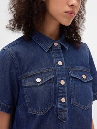 Chemise en denim avec un col, manches courtes, deux poches poitrine et boutons en ton cuivre ; tissu bleu foncé avec une finition texturée.