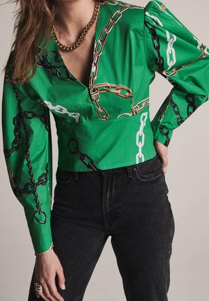 Femme portant un chemisier vert avec un motif chaîne multicolore, des manches bouffantes, un jean noir taille haute et un collier chaîne en or.