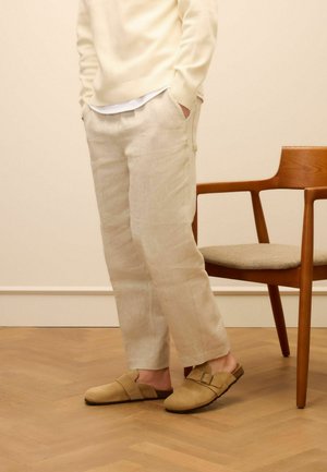 Beige linen trousers with a relaxed fit, paired with brown suede slip-on shoes featuring a cross strap and buckle accent.