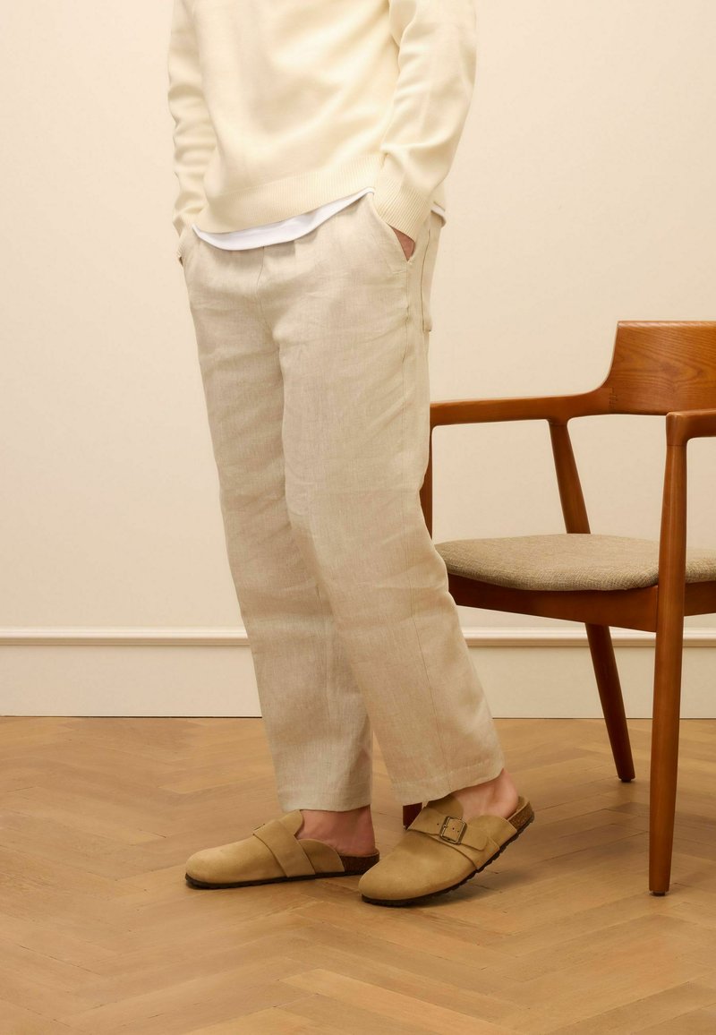 Beige linnen broek met een relaxte pasvorm, gecombineerd met bruine suède instappers met een kruislint en een gespaccent.}}