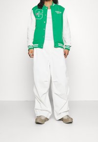 Chaqueta varsity verde y blanca con un gran emblema de "C", con puños y dobladillo a rayas. Combinada con pantalones blancos de corte holgado y zapatos beige.