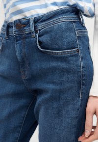 Κοντινό πλάνο μπλε τζιν με μπροστινές τσέπες και λεπτομέρεια κουμπιών, φορεμένο με λευκό μπλουζάκι με μπλε ριγέ μοτίβο που είναι τυλιγμένο μέσα στο τζιν.