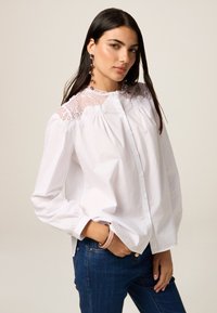 Camicia bianca con bottoni, dettagli in pizzo sulle spalle, maniche lunghe, tessuto arricciato e morbido, abbinata a pantaloni di jeans.