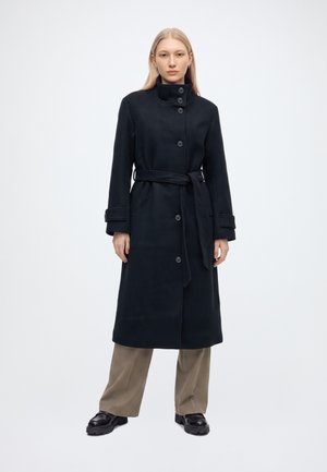 Femme aux cheveux raides et clairs portant un long manteau noir ceinturé, un pantalon beige à jambes larges et des chaussures noires épaisses, debout devant un fond uni.