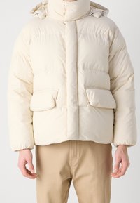 Puffjakke i off-white med høj krave, frontknapper, to store lommer og elastiske manchetter. Blød, quilted tekstur.