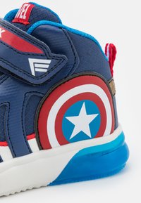 Shoe desportivo azul com detalhes em vermelho e branco, apresentando um emblema redondo com uma estrela, material texturizado e uma sola grossa e transparente.
