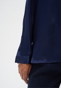 Granatowa satynowa koszula z długim rękawem, o gładkiej, błyszczącej fakturze z mankietem i subtelnym rozcięciem przy nadgarstku, trzymana w dłoni.