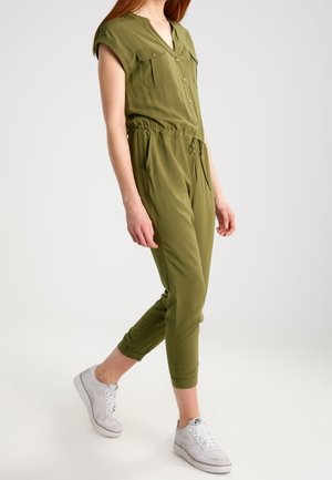 Vrouw die loopt en een olijfgroene korte mouwen jumpsuit met zakken en een taillekoord draagt, gecombineerd met witte sneakers.