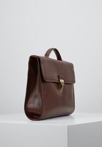 Serviette en cuir marron avec poignée supérieure et fermeture à loquet doré, debout sur une surface blanche devant un fond gris.
