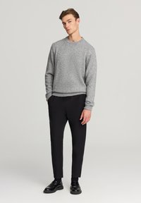 Maglione grigio a maglia con scollatura rotonda, abbinato a pantaloni neri tapered e scarpe nere, su uno sfondo neutro.