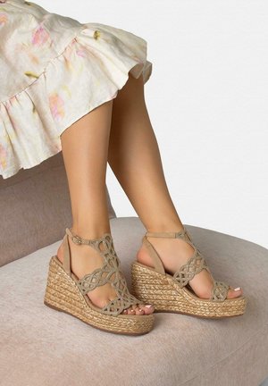 Sandalias de cuña tejidas con tiras recortadas intrincadas y hebilla en el tobillo, usadas con un vestido de dobladillo con volantes florales, sentada en un otomán beige suave.