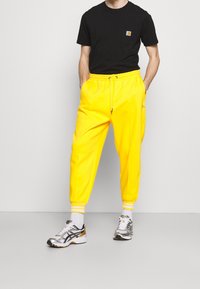 Schwarzes T-Shirt mit kleinem Logo auf der Brust, kombiniert mit leuchtend gelben Jogginghosen mit weißen gestreiften Bündchen und einem elastischen Bund.