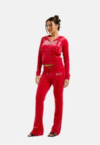 Tuta in velluto rosso composta da una giacca a zip cropped e pantaloni a zampa, con la scritta "JUICY" in strass. Tessuto morbido e design aderente.