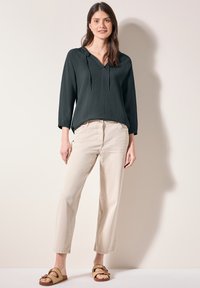 Top lungo a maniche lunghe di un verde scuro, texture, con vestibilità morbida e scollo a V, abbinato a pantaloni beige chiaro a gamba dritta e sandali intrecciati.