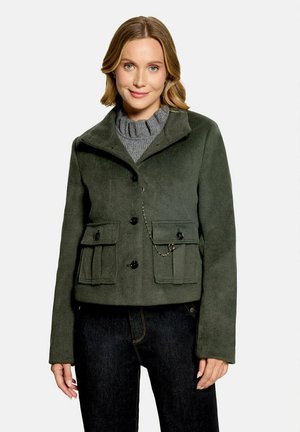 Femme portant une veste boutonnée vert foncé avec poches à rabat et détail de chaîne argentée sur un pull en maille gris et un pantalon foncé, souriant légèrement.