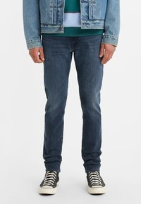 Denim jeans in dark blue, slim fit, featuring a smooth texture. Paired with black high-top sneakers and a light blue denim jacket.