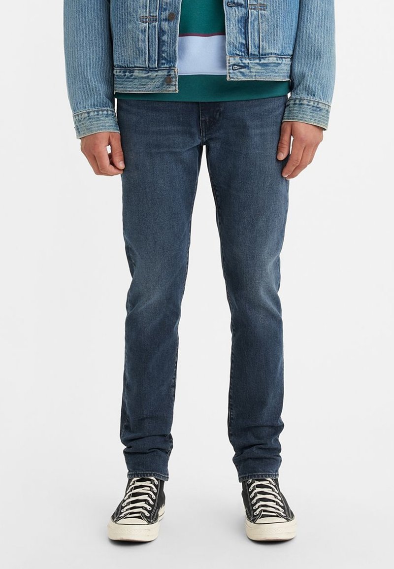 Denim jeans in dark blue, slim fit, featuring a smooth texture. Paired with black high-top sneakers and a light blue denim jacket.