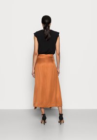 Nainen, jolla on tummat hiukset poninhännällä, pukeutuneena mustaan hihattomaan toppiin, poltetuista oransseista midi-hameeseen ja mustiin korkoihin, seisoo selkä kameraan päin.