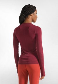 Bordeauxfarbendes Langarmoberteil aus einem glatten, dehnbaren Stoff mit einer figurnahen Passform; getragen mit kontrastierenden roten Leggings.