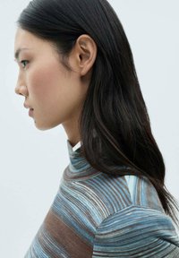 Turtlenecktröja med blå och bruna horisontella ränder, ribbat mönster och hög krage. Håret faller löst förbi axlarna.