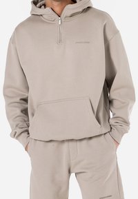 Sweat à capuche beige zippé avec une poche kangourou, doté d'une texture douce et de poignets côtelés. Logo imprimé sur la poitrine.