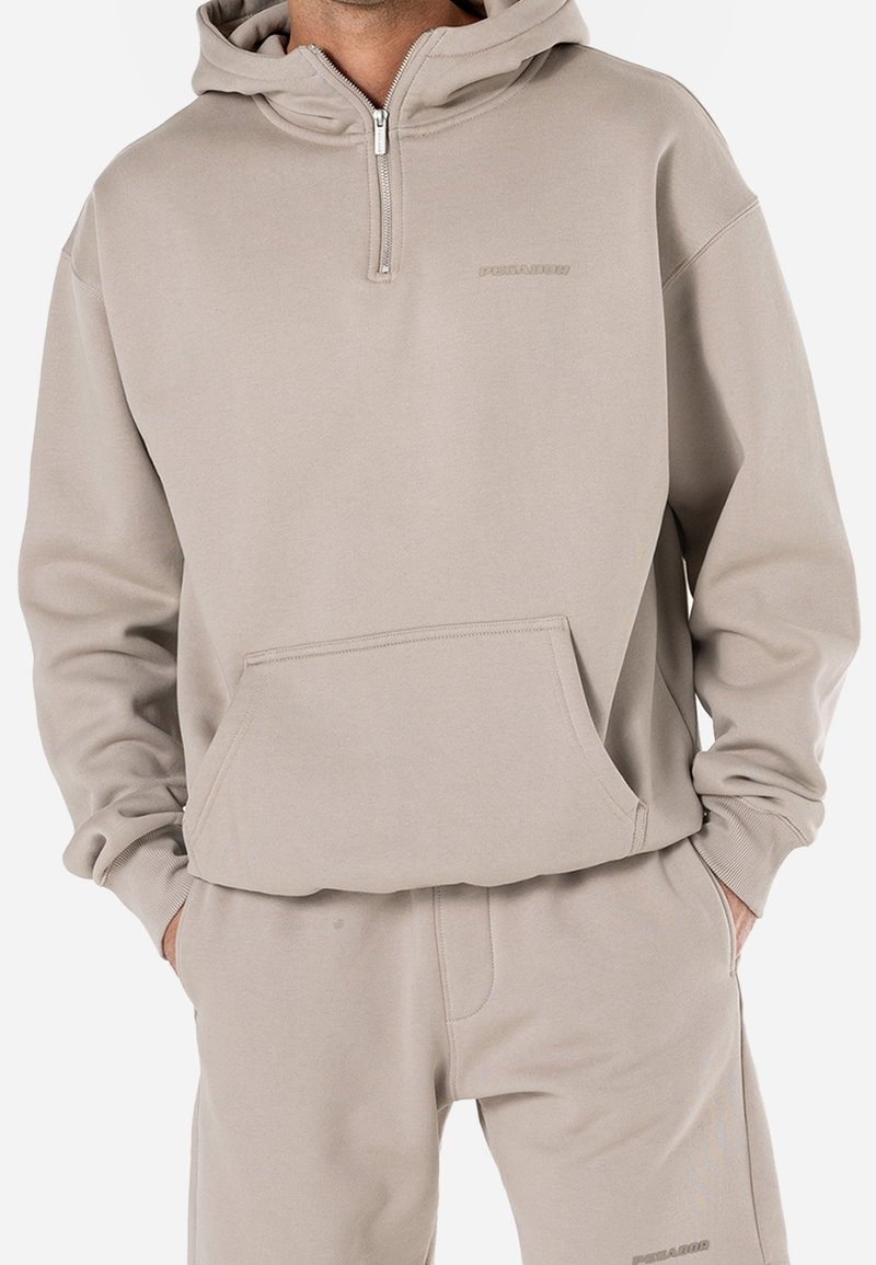 Sweat à capuche beige zippé avec une poche kangourou, doté d'une texture douce et de poignets côtelés. Logo imprimé sur la poitrine.