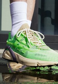 Grüne Netzsport-Schuhe mit beigen Akzenten, Schnürsenkeln und einer strukturierten Sohle. Sichtbare Reflexion im Wasser. Weiße Crew-Socke über dem Knöchel.