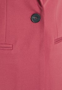 Růžový strukturovaný blejzr s jedním černým knoflíkem a dvěma klopovými kapsami. Tkanina má jemný žebrovaný vzor.