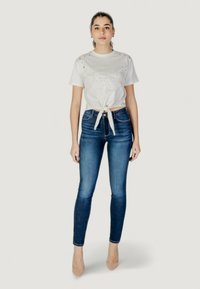 White cropped t-shirt with a knot detail and gold embellishments, paired with fitted dark blue jeans and nude high heels.