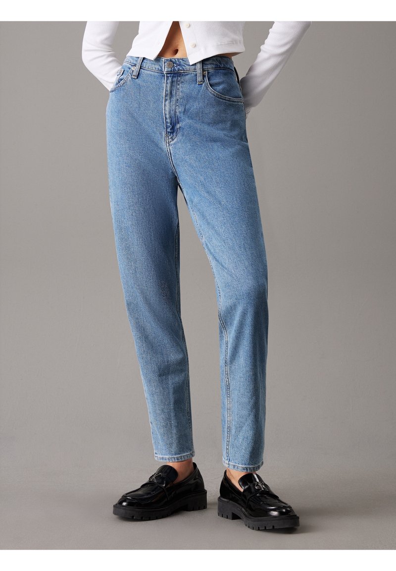 Person som bär högmidjade ljusblå raka jeans med framfickor och svarta kraftiga loafers på ett grått golv.