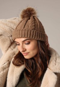 Bruine gebreide beanie met een textuurpatroon en een pluche pompon bovenop, gedragen met een crèmekleurige nepbontjas. Zacht, warm materiaal.